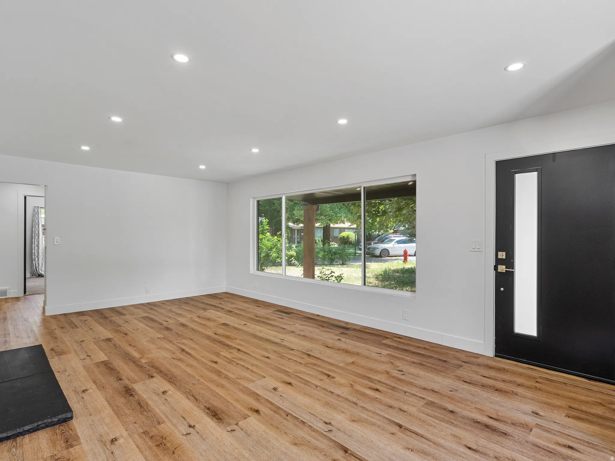 Spacious living area featuring wood-finish flooring, recessed ceiling lighting, a large picture window, and a modern entry door with a vertical frosted glass insert