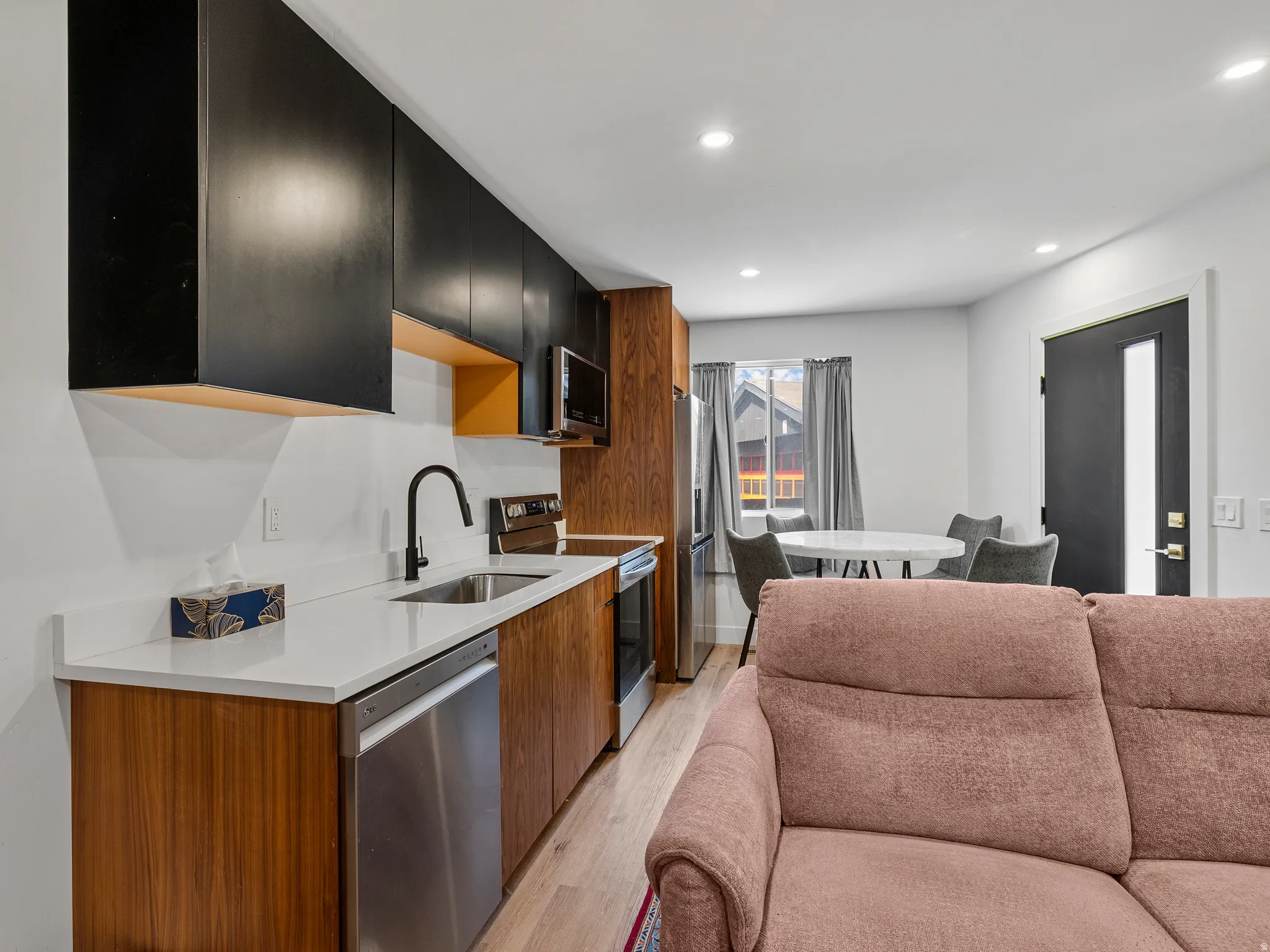 Modern kitchen featuring two-tone cabinetry with black upper cabinets and wood-finish lower cabinets, stainless steel appliances, a single-basin sink with a matte black faucet, and light-colored countertops