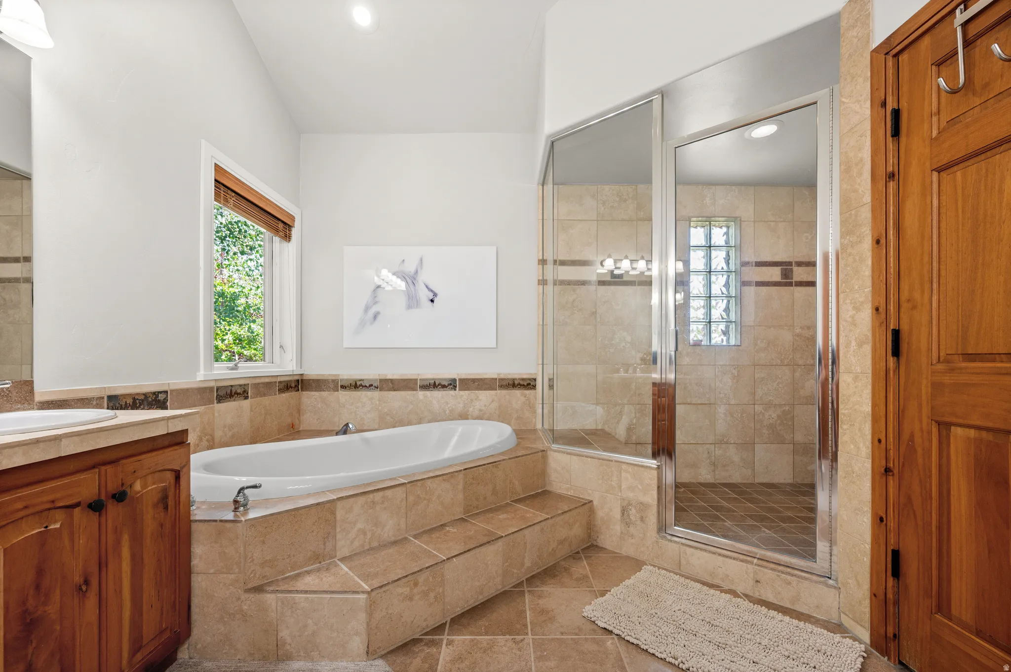 Spacious bathroom featuring a built-in soaking tub with tiled surround, a separate glass-enclosed shower with tiled walls and a glass block window, and dual vanities with wood cabinetry and undermount sinks