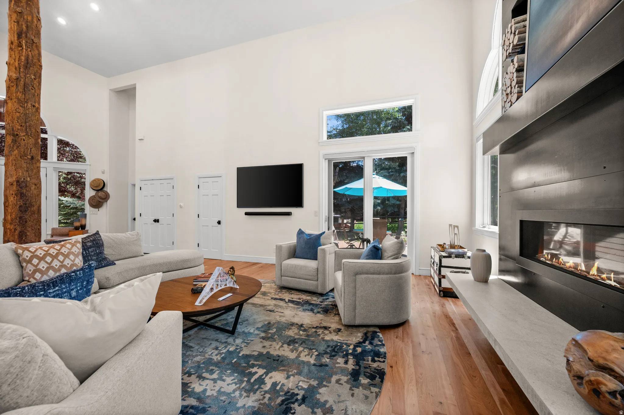 Spacious living area featuring high ceilings, hardwood flooring, a prominent tree-trunk architectural feature, a contemporary fireplace with a wide hearth, and glass French doors leading to exterior grounds