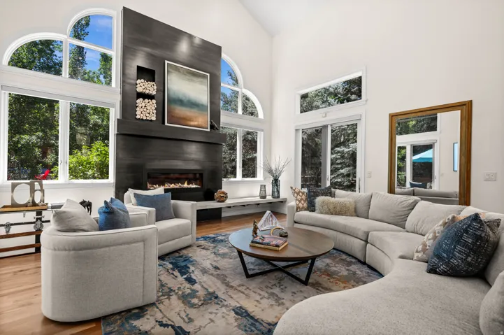 Two-story living room featuring a tall, dark-paneled fireplace, vaulted ceilings, natural wood-finish flooring, and large arched windows