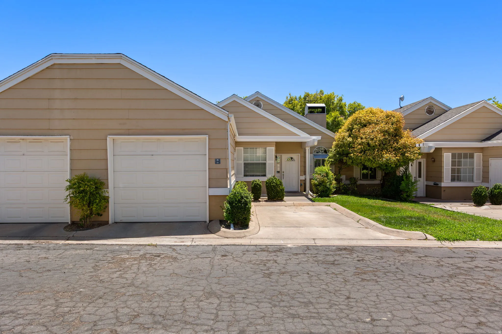 Exterior featuring tan siding, white trim, two white garage doors, a paved driveway, and mature landscaping