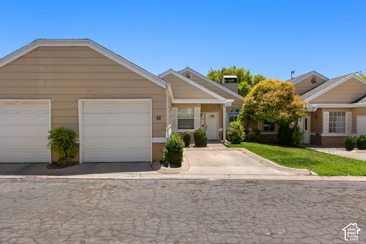 Exterior featuring tan siding, white trim, two white garage doors, a paved driveway, and mature landscaping
