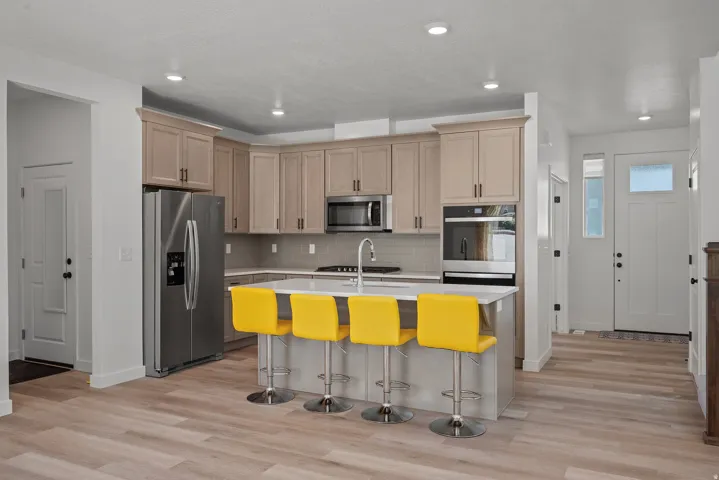 Contemporary kitchen featuring light wood-finish cabinetry, stainless steel appliances, a white island with an integrated sink, and wood-finish flooring extending into the entry