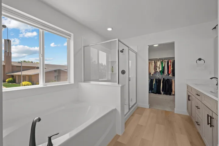 Bathroom featuring a built-in soaking tub, a glass-enclosed shower, a double vanity with light-toned cabinetry and stone-finish countertops, and wood-finish flooring