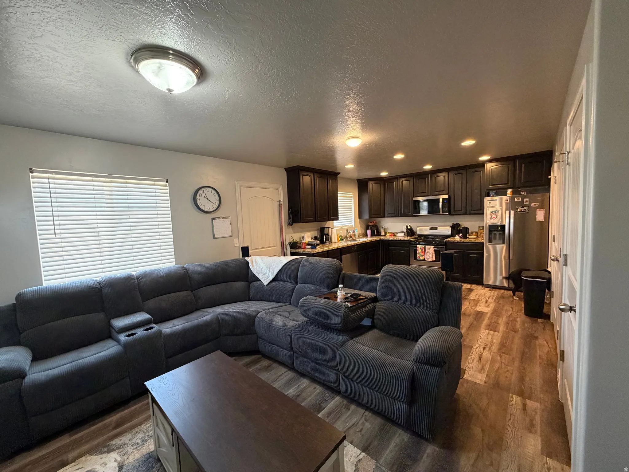 Open-concept living space featuring wood-finish flooring, dark wood cabinetry, stainless steel appliances, and recessed lighting