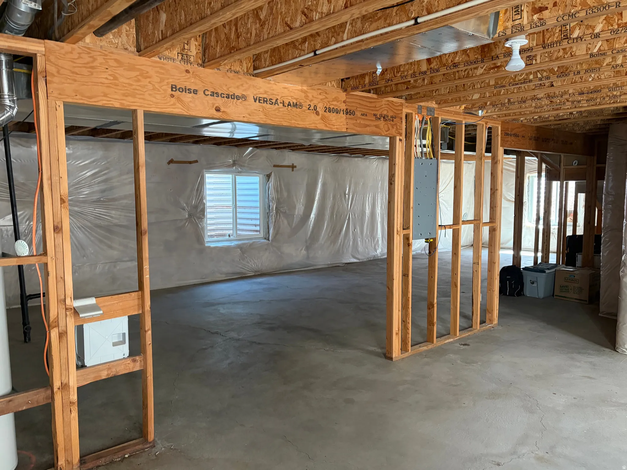 Unfinished basement space featuring exposed wooden joists and framing, concrete flooring, window with louvered blinds, and visible electrical panels