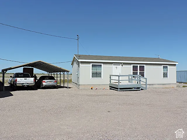 Light grey exterior with white trim, featuring a front porch with railing, a two-car metal carport, and a gravel lot