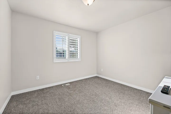 Bright room featuring a window with white plantation shutters, neutral-toned walls, and grey carpet flooring