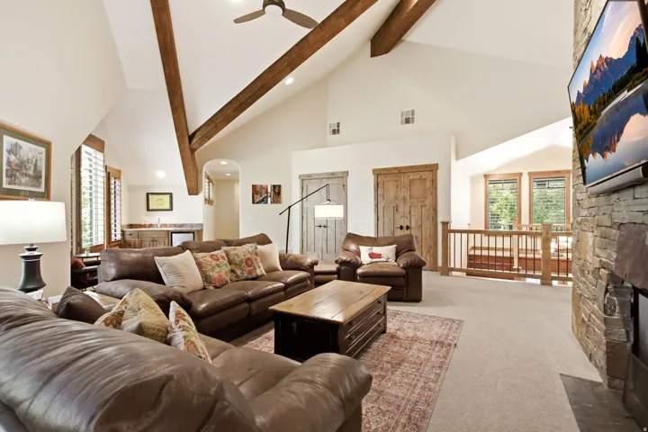 Vaulted ceilings with exposed wood beams, a stone-clad fireplace, and a built-in wet bar with cabinetry define this expansive living area