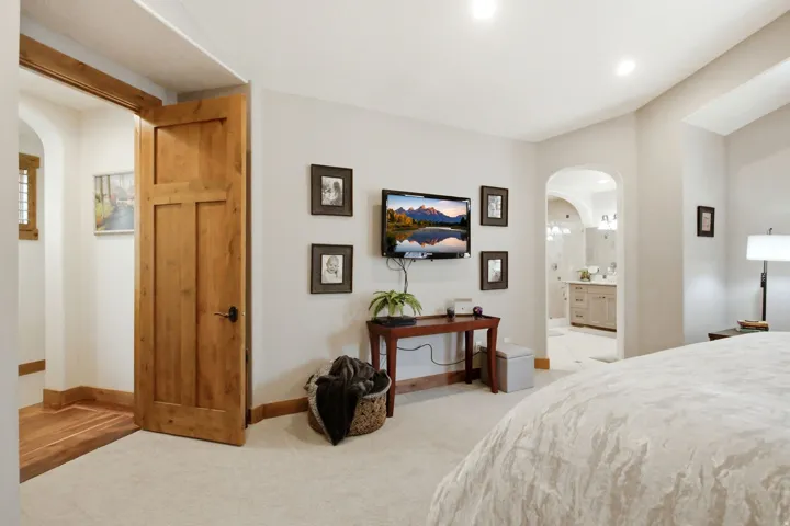 Carpeted sleeping area featuring a wood-panel door, recessed lighting, and an arched entry to a bathroom with a double vanity and sconce lighting