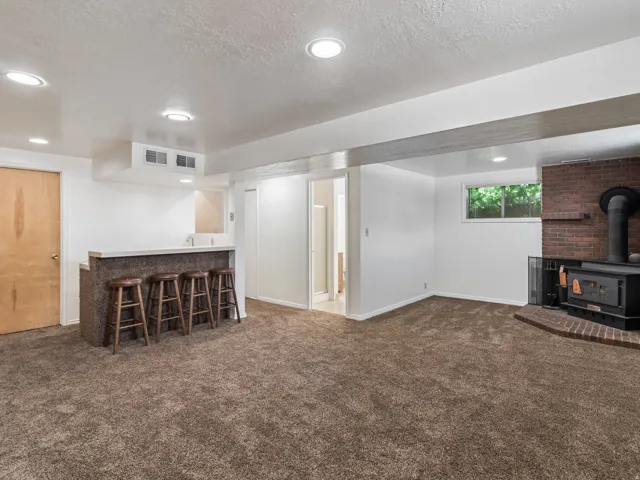 Spacious room featuring a built-in wet bar with a countertop and sink, a brick fireplace with a wood-burning stove, brown carpet flooring, and recessed ceiling lighting