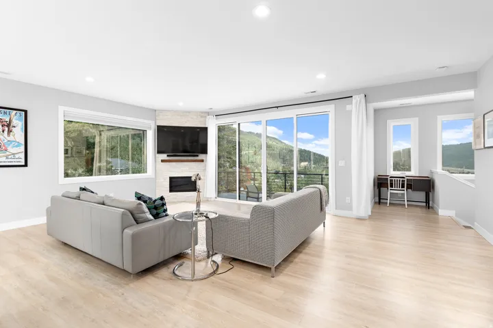 Spacious living area featuring light wood-finish flooring, a corner fireplace with a stacked stone facade and wood mantel, large sliding glass doors opening to a balcony, and abundant natural light from multiple windows