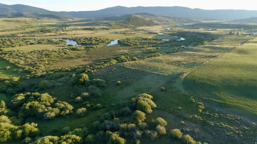Expansive rural landscape featuring a winding river, multiple ponds, lush green fields, and distant mountain ranges