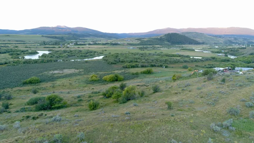 Expansive landscape featuring rolling hills, abundant green foliage, a distant body of water, and mountainous terrain under a clear sky