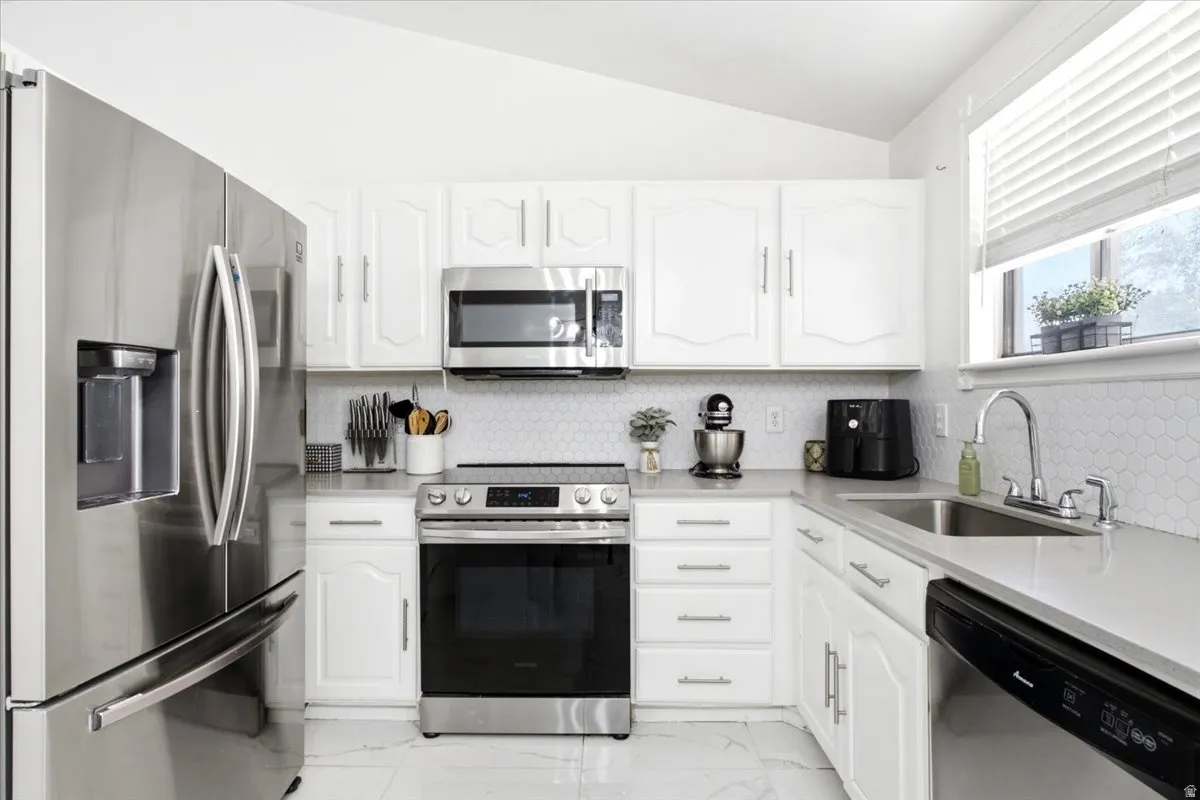 Kitchen featuring white cabinetry, light-toned countertops, a hexagonal tile backsplash, stainless steel appliances, and a window with horizontal blinds