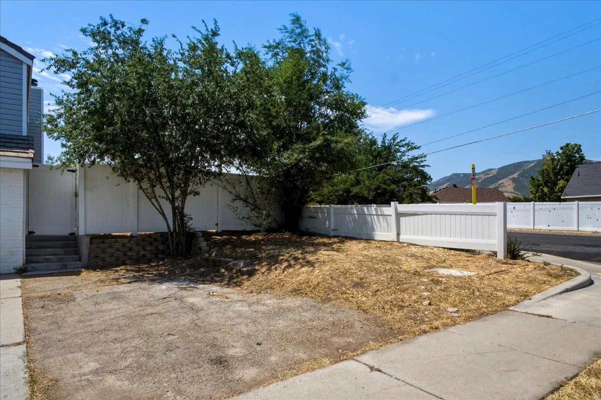 Front yard space featuring a white privacy fence, mature trees, retaining wall, and concrete pad