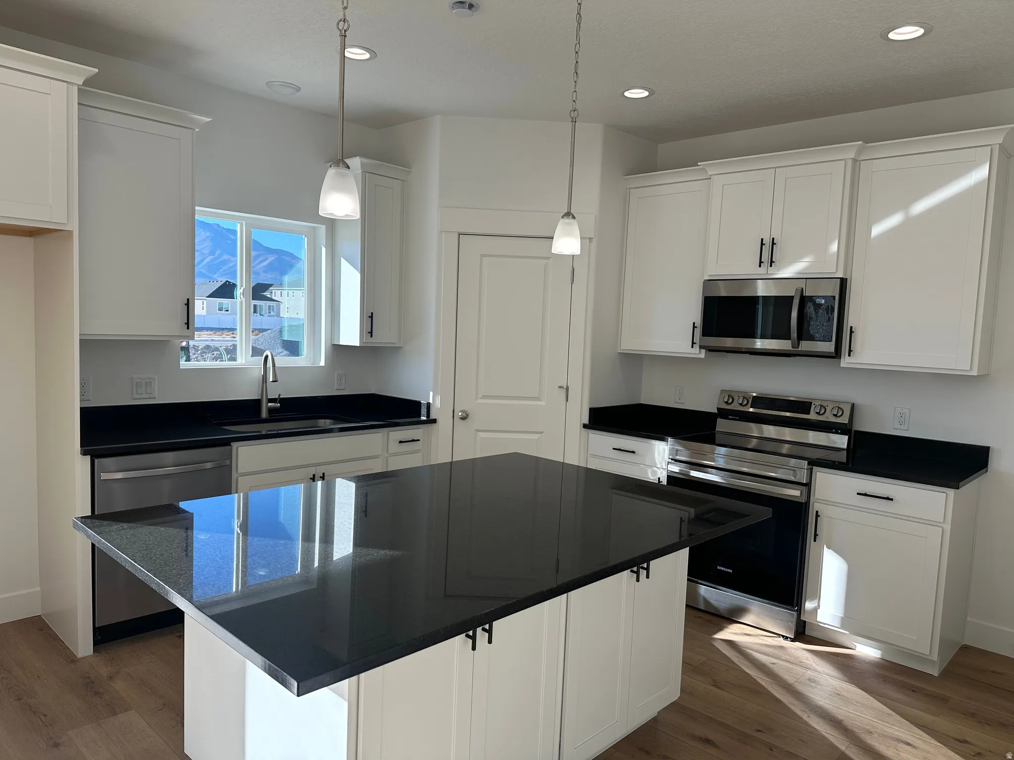 Modern kitchen featuring white shaker-style cabinetry, dark countertops, wood-finish flooring, stainless steel appliances, and a central island with pendant lighting