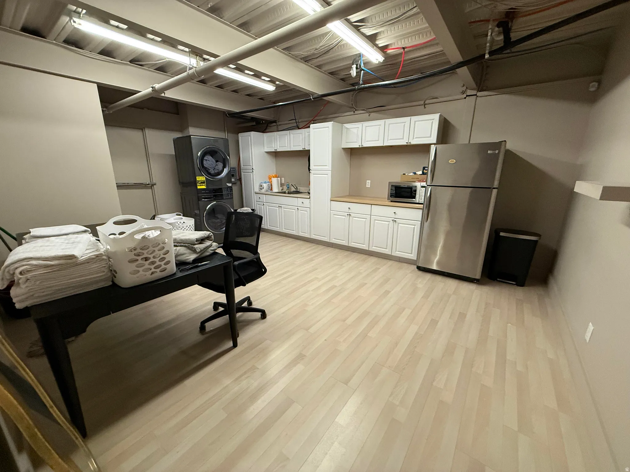 Spacious utility area featuring light wood-finish flooring, white cabinetry, a stainless steel refrigerator, and stacked laundry machines