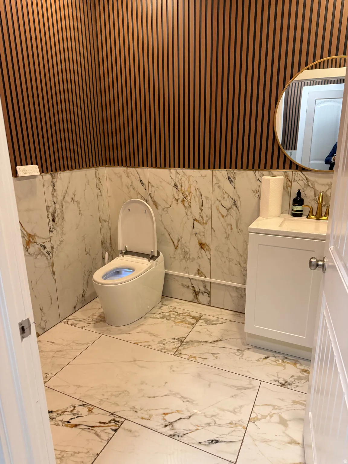 Powder room featuring wood-slat wall paneling, full-height marble-look tile, and a circular wall mirror with a gold-finish frame