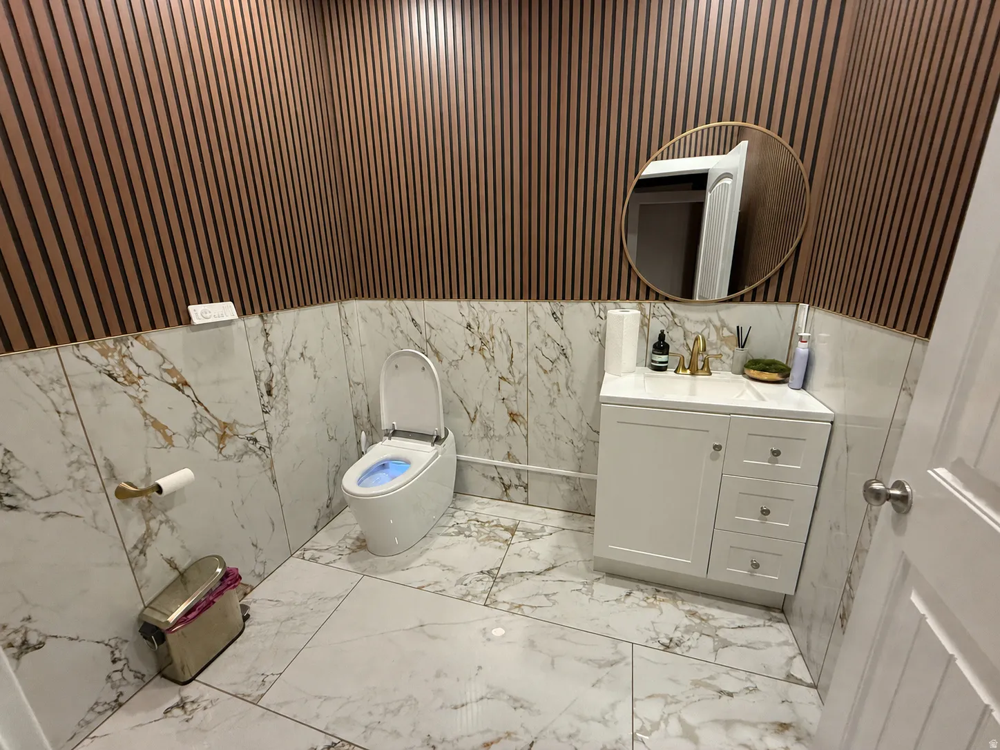 Powder room featuring marble-look wall and floor tiling, wood-slat accent wall, white vanity with gold-tone faucet, and a round gold-framed mirror