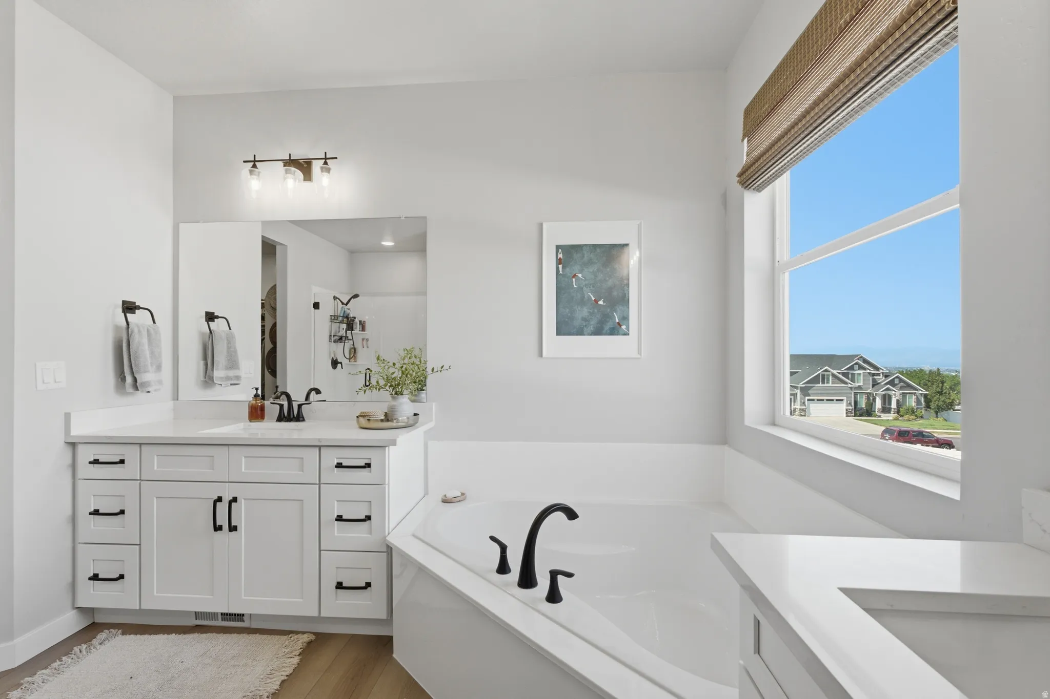Ensuite Bathroom featuring a built-in corner soaking tub with a black-finish faucet, a large window with a woven shade, and a vanity with white cabinetry.