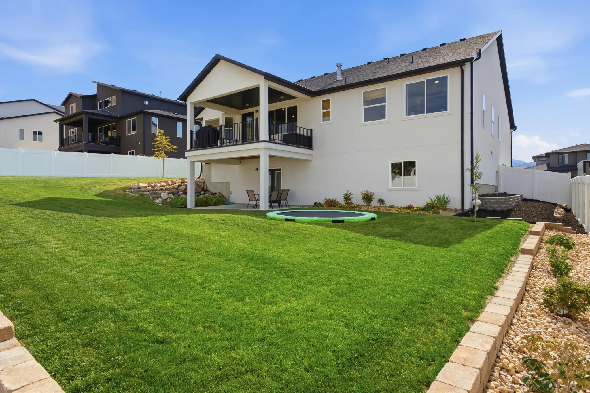 Spacious backyard with a vibrant green lawn, modern white siding exterior, covered upper-level deck with black railing, lower-level patio, and retaining wall landscaping