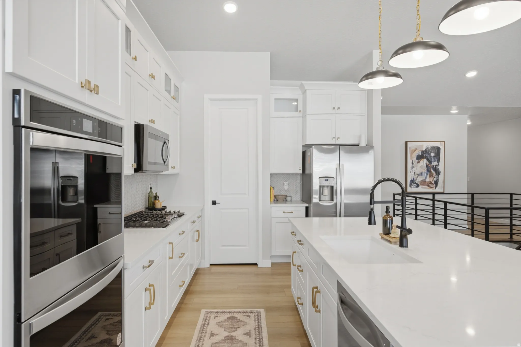 Modern kitchen featuring white cabinetry with gold-tone hardware, a spacious island with an undermount sink and matte black faucet, stainless steel appliances including a double oven and refrigerator, and light wood-finish flooring