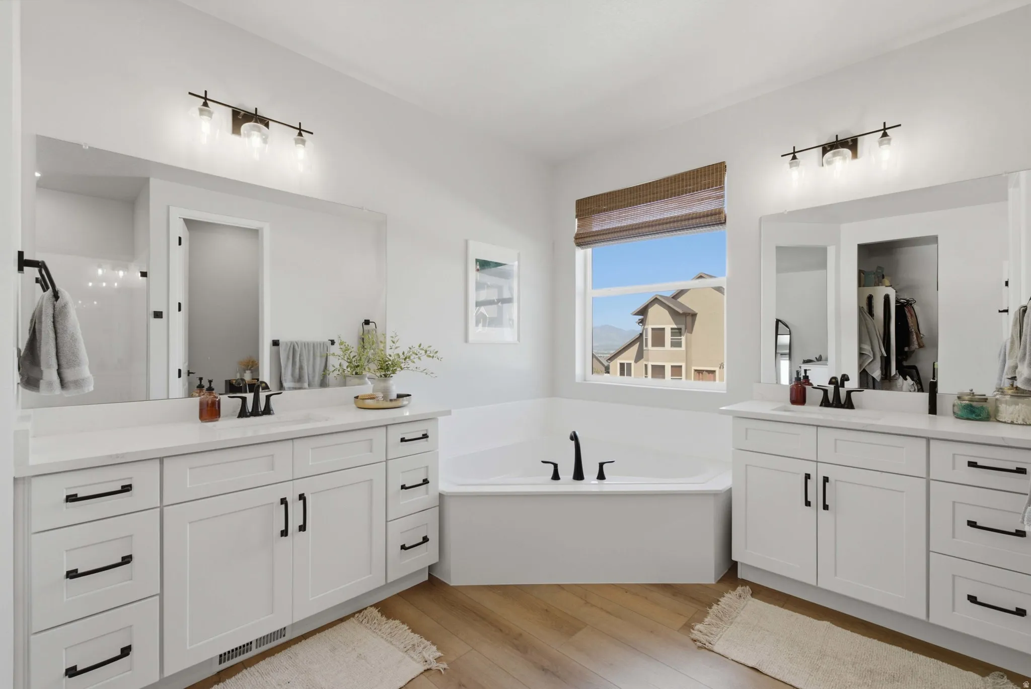 Ensuite Bathroom featuring a built-in soaking tub with black fixtures, dual white shaker vanities with quartz countertops, wood-finish flooring, and a large window with a woven shade