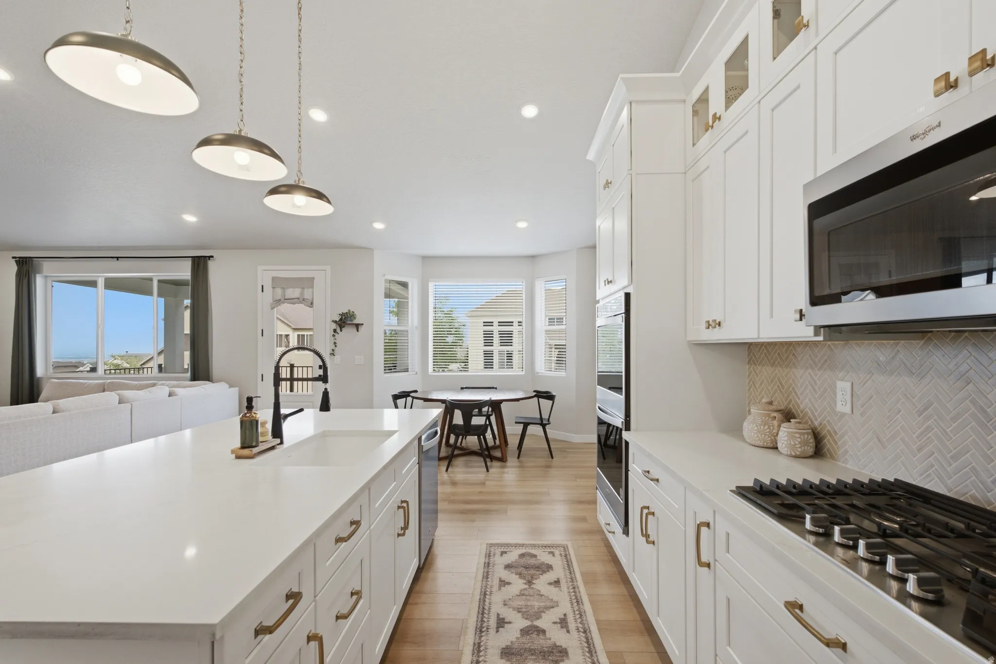 Gourmet kitchen featuring a large island with an undermount sink, white cabinetry with brass hardware, a gas cooktop, double wall ovens, and a herringbone tile backsplash