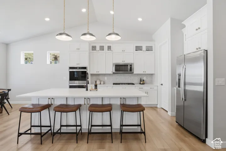 Modern kitchen featuring a large central island with seating, stainless steel appliances, white cabinetry with glass-front accents, and wood-finish flooring