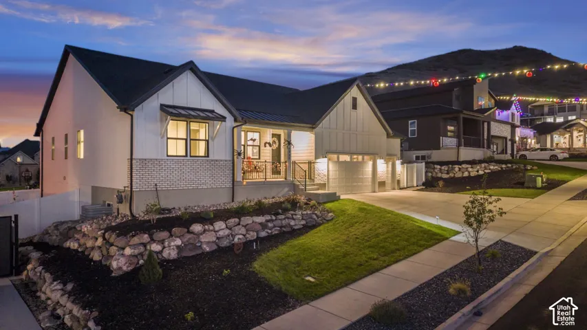 Modern farmhouse-style exterior featuring a covered porch with metal railing, an attached garage, a stone retaining wall, and a manicured lawn