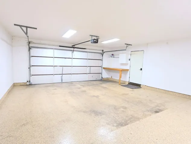 Spacious two-car garage featuring an epoxy-coated concrete floor, two rectangular overhead light fixtures, and a built-in workbench with an upper cabinet