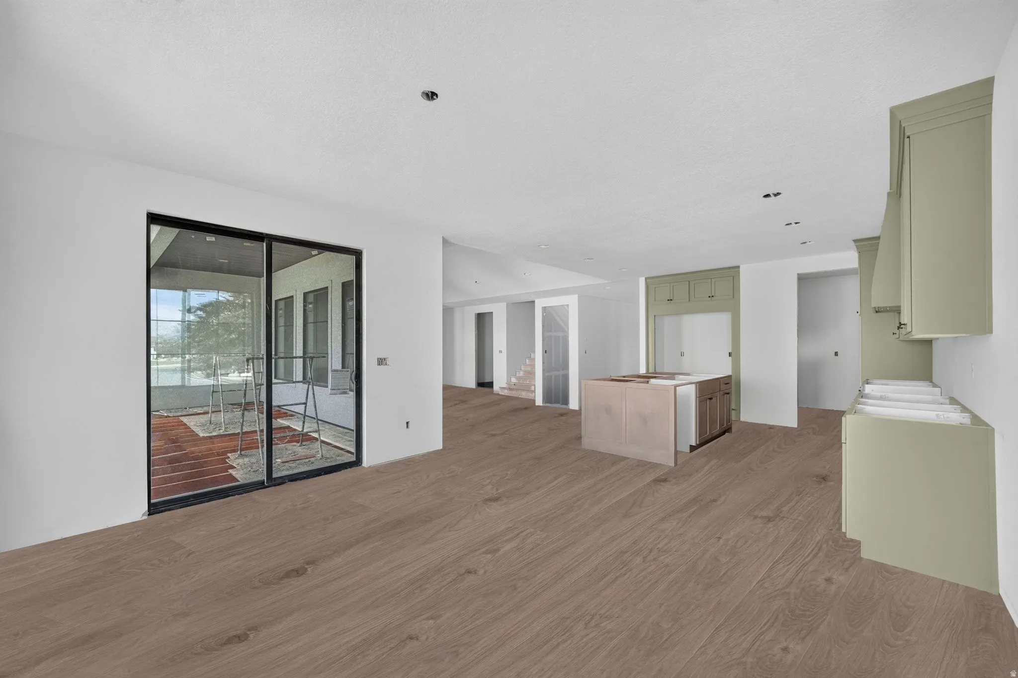 Expansive open-concept space featuring wood-finish flooring, a kitchen island with built-in cabinetry, sage green cabinetry, recessed lighting, and large sliding glass doors leading to a covered outdoor area