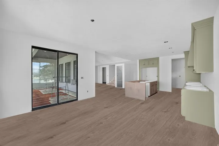 Expansive open-concept space featuring wood-finish flooring, a kitchen island with built-in cabinetry, sage green cabinetry, recessed lighting, and large sliding glass doors leading to a covered outdoor area