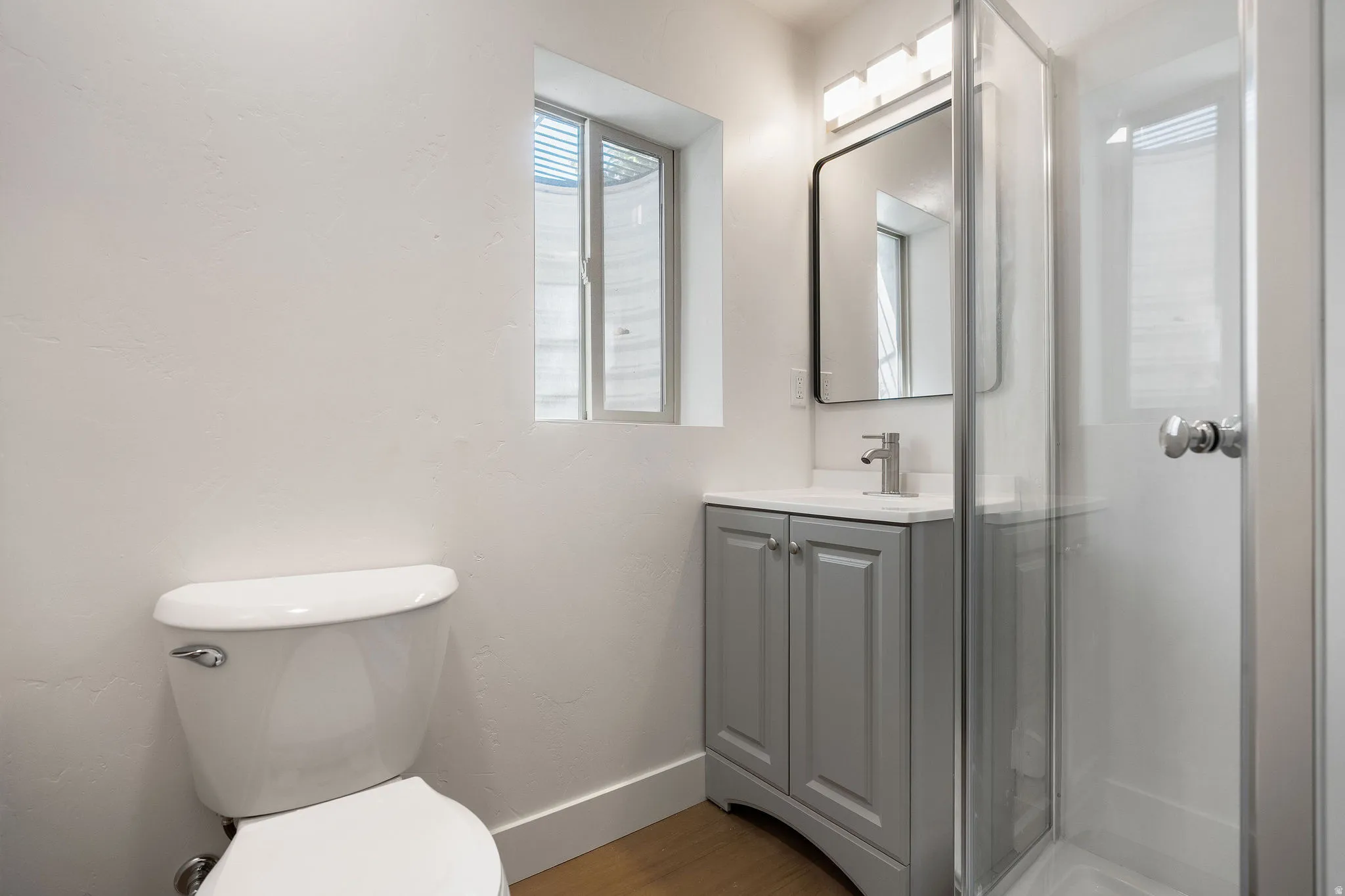 Bathroom featuring a glass-enclosed shower, a gray vanity with a white countertop and chrome faucet, a rectangular mirror, and a modern light fixture