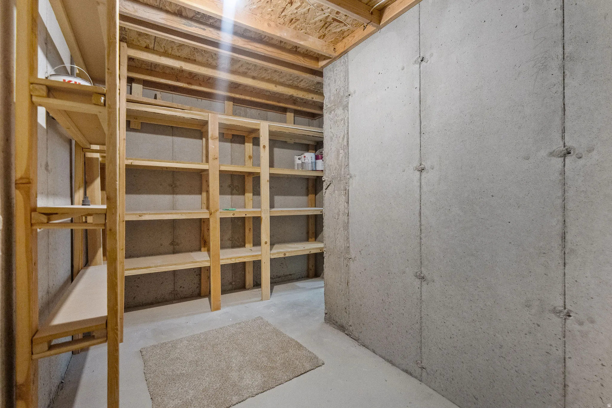 Basement storage area featuring extensive built-in wooden shelving, exposed ceiling joists, concrete walls, and a light-colored concrete floor