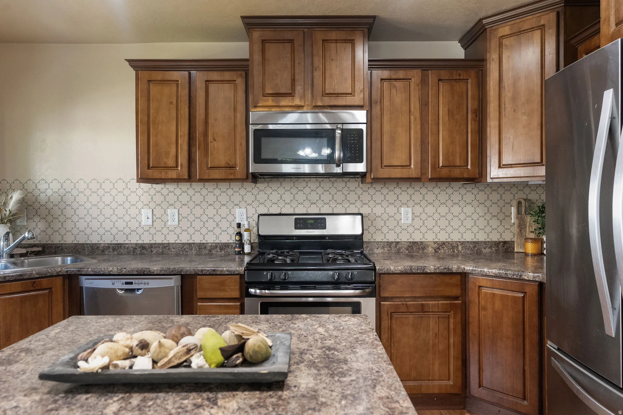 Kitchen featuring wood cabinetry, stainless steel appliances, patterned tile backsplash, and a central island with a stone-finish countertop