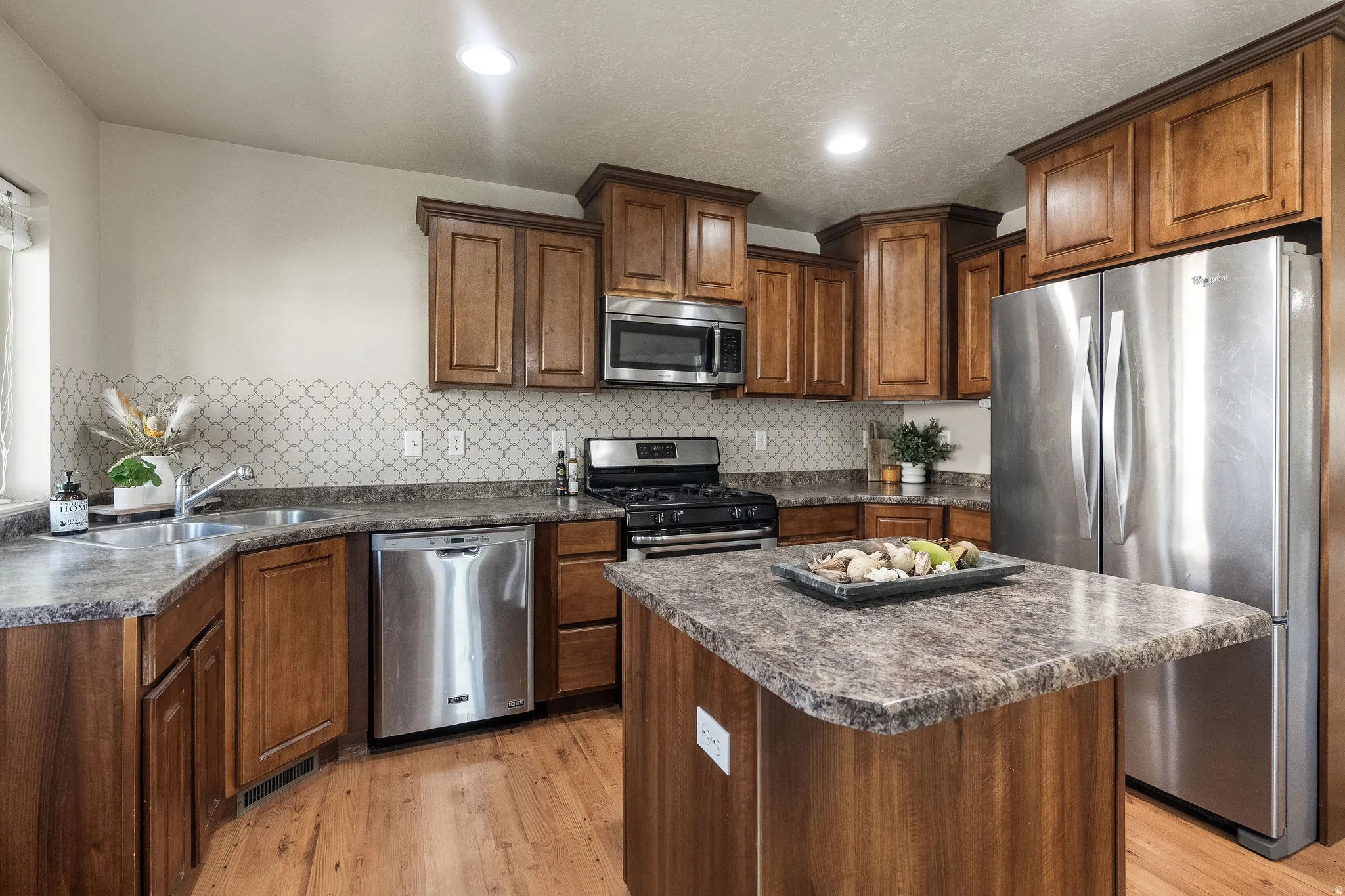 Kitchen featuring wood-finish cabinetry, stainless steel appliances, a patterned tile backsplash, wood-finish flooring, and a central island with countertop space
