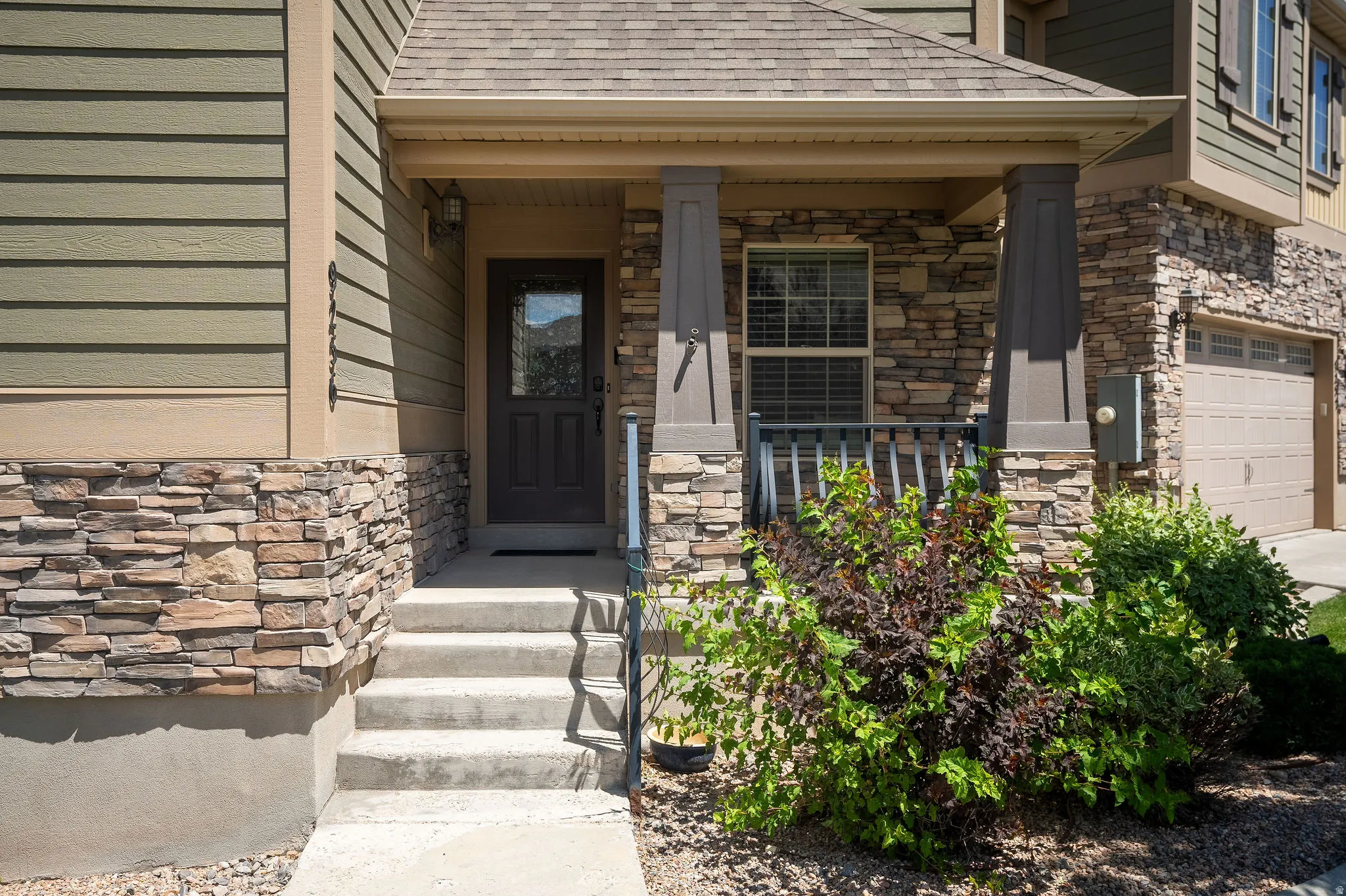 Covered entryway featuring stacked stone accents, horizontal siding, concrete steps with metal railing, an integrated garage, and established landscaping