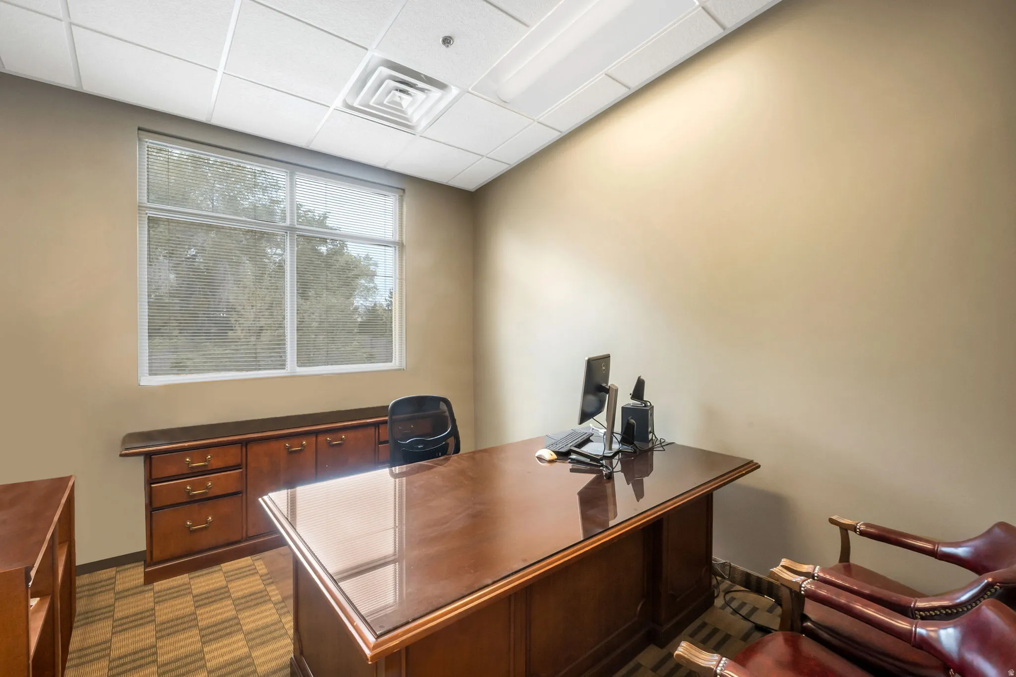Office space featuring a large window with horizontal blinds, neutral wall tones, and ceiling grid with integrated lighting