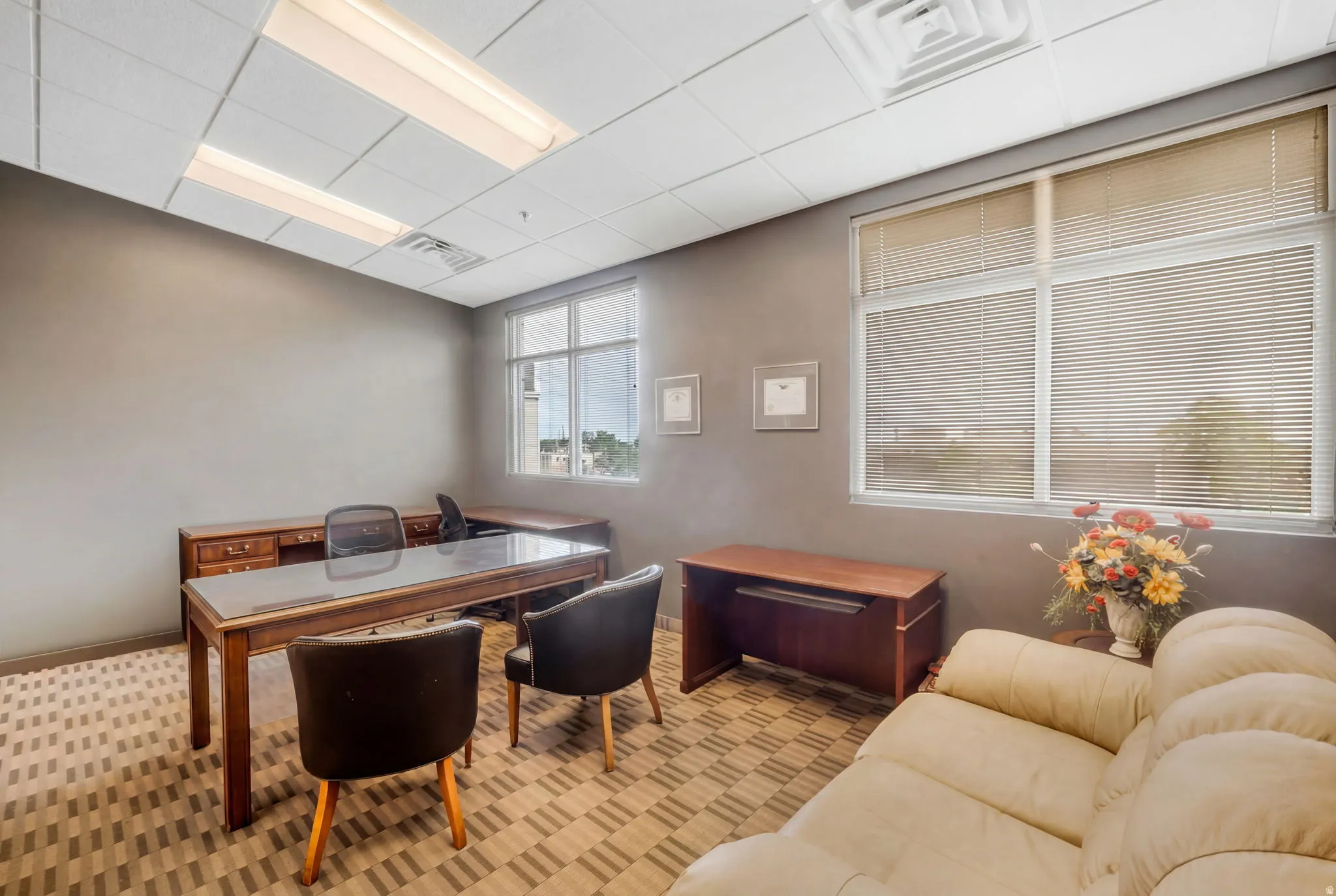 Office space featuring a suspended acoustic ceiling with integrated fluorescent lighting, patterned carpet flooring, and multiple windows with horizontal blinds