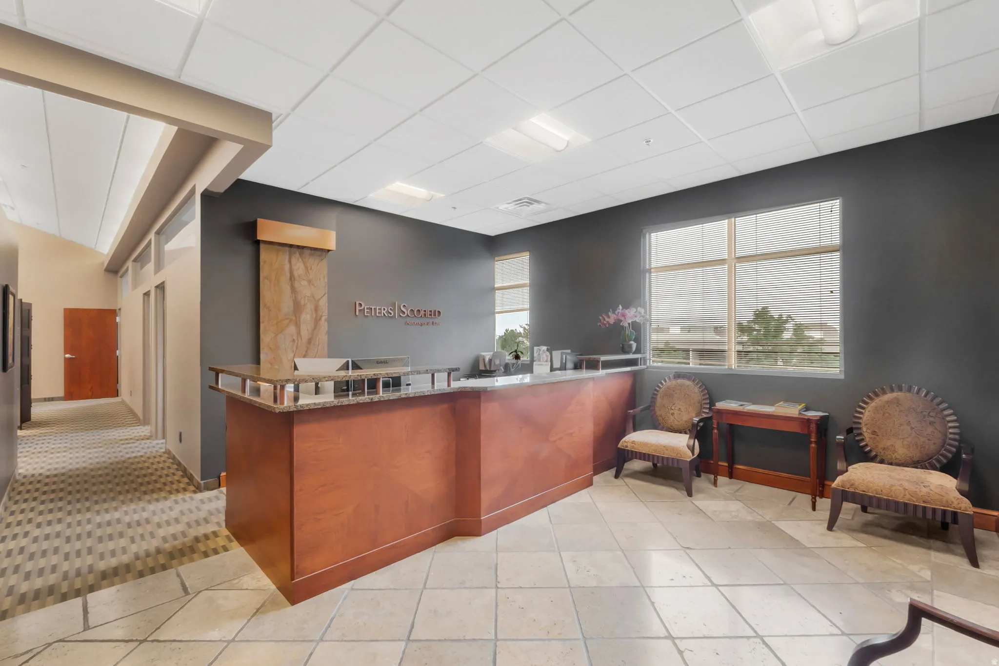 Spacious reception area featuring a wood-finish reception desk with a stone countertop, an integrated water feature, large windows with horizontal blinds, and tile flooring