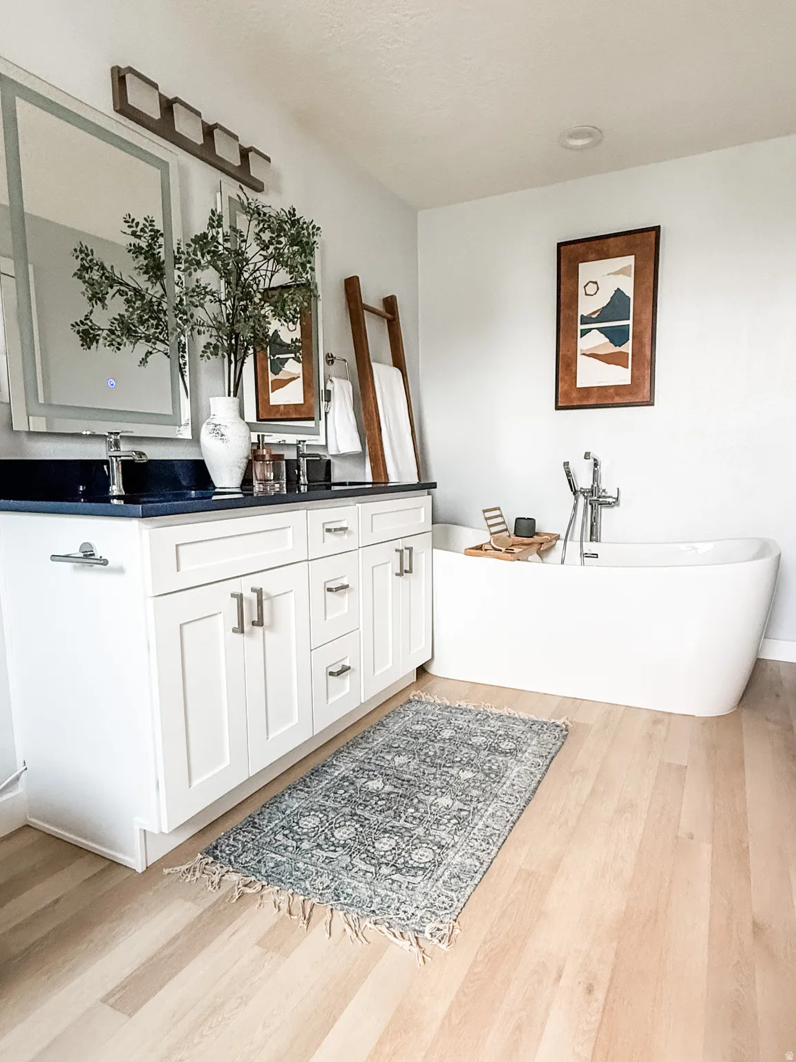 Bathroom featuring a freestanding soaking tub with floor-mounted faucet, a double vanity with white shaker-style cabinetry, dark countertops, and dual illuminated mirrors