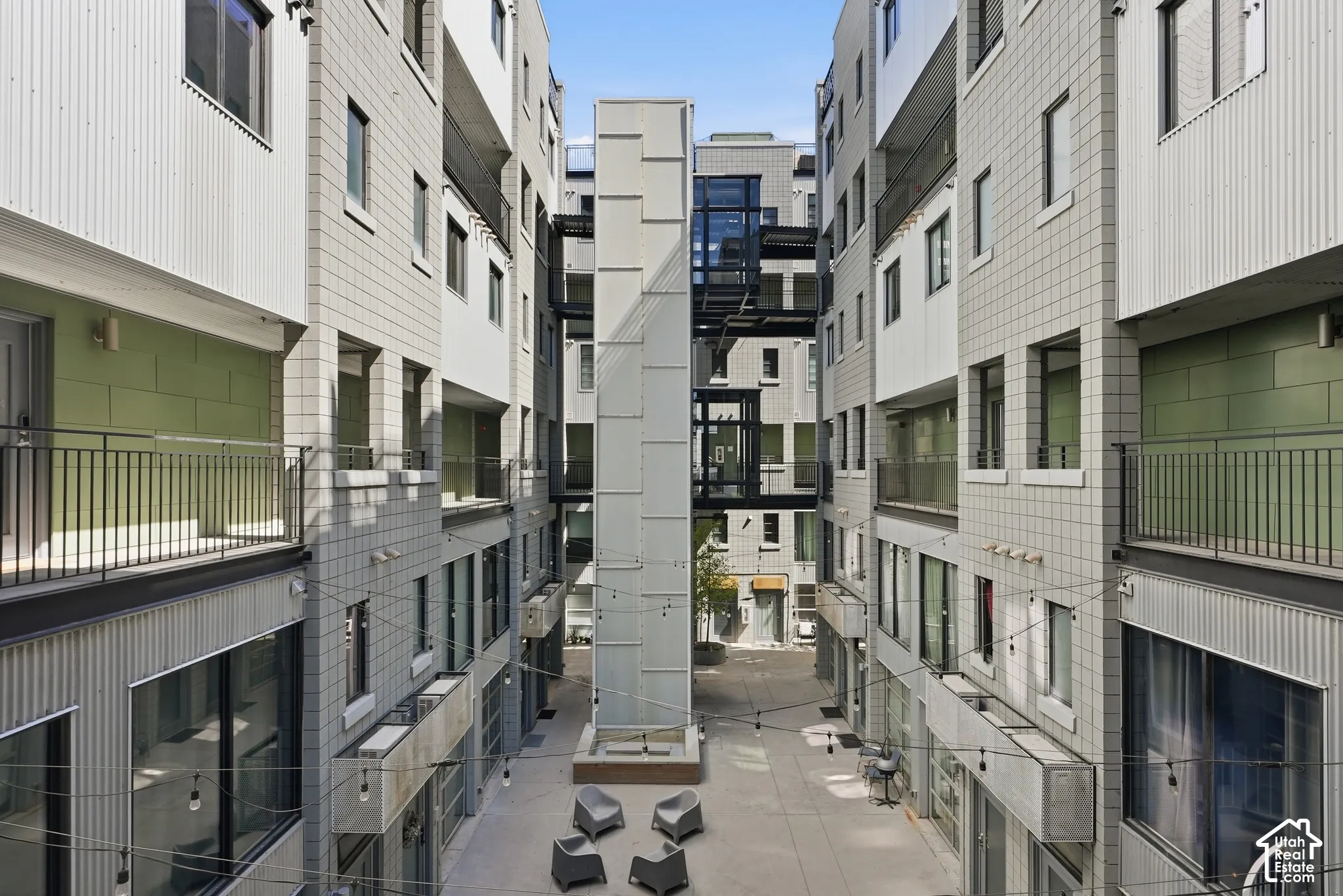 Modern multi-level building exterior featuring corrugated metal and square-paneled siding, multiple balconies with dark railings, a central glass-enclosed elevator shaft, and an open-air courtyard with seating