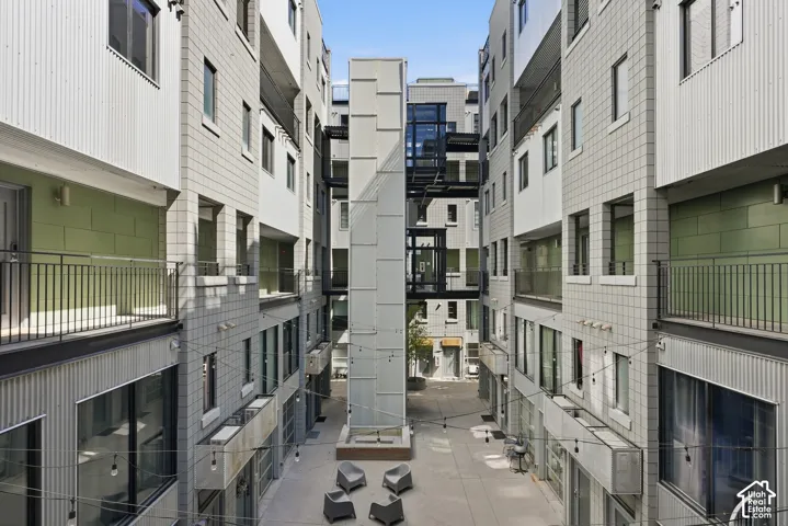 Modern multi-level building exterior featuring corrugated metal and square-paneled siding, multiple balconies with dark railings, a central glass-enclosed elevator shaft, and an open-air courtyard with seating