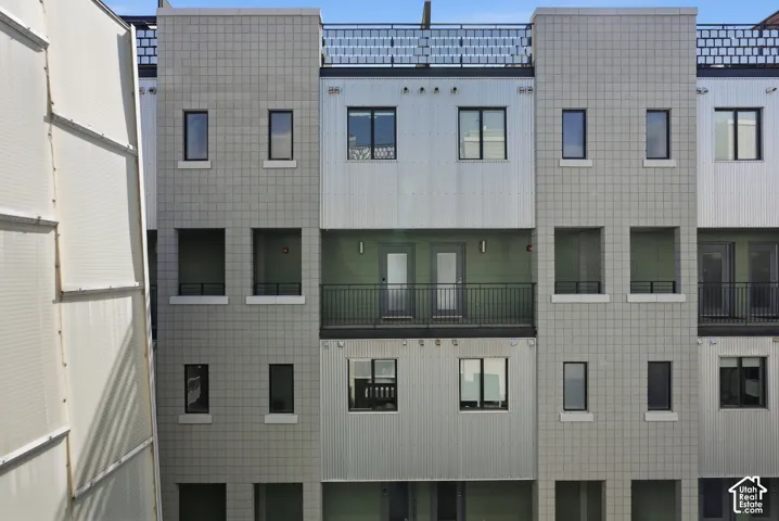Multi-level contemporary facade featuring a central balcony with dark metal railings, contrasting smooth and corrugated paneling, and multiple dark-framed windows