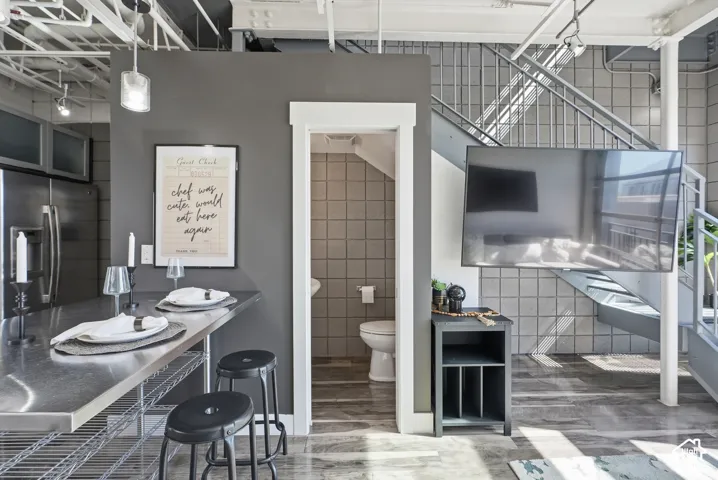 Open-concept living space featuring a stainless steel kitchen island with seating, exposed industrial-style ceiling beams, a contemporary open staircase, and wood-finish flooring