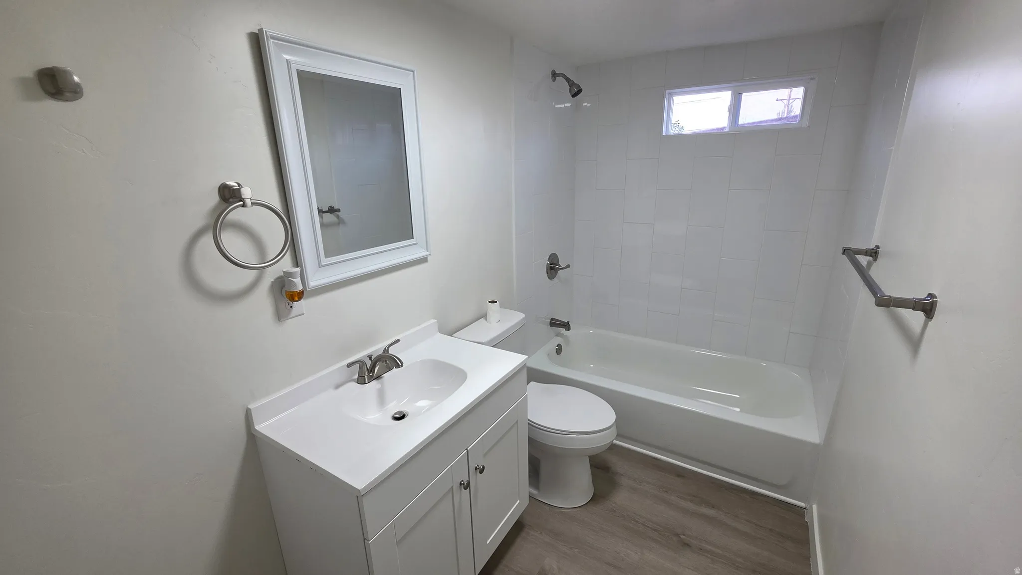 Bathroom featuring a white vanity with an integrated sink, a framed mirror, and a white tiled shower/tub combination with a high window