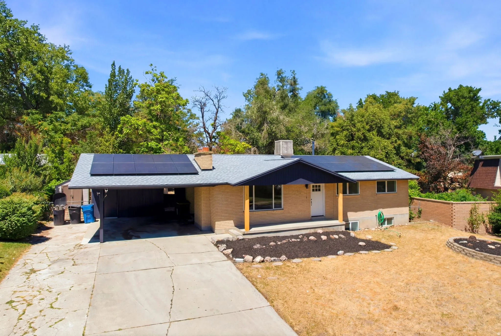 Ranch-style residence featuring a brick facade, carport, solar panel array, front porch, and a tiered front yard with landscape beds